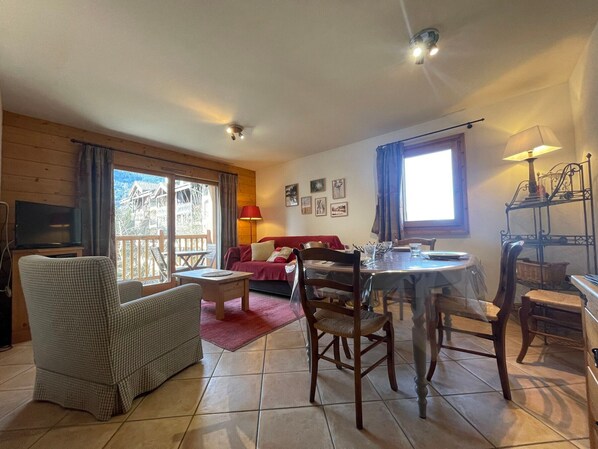 Een gezellige woonkamer met een houten vloer, een rode bank, een eettafel met stoelen en een balkon met uitzicht op een bergchalet.