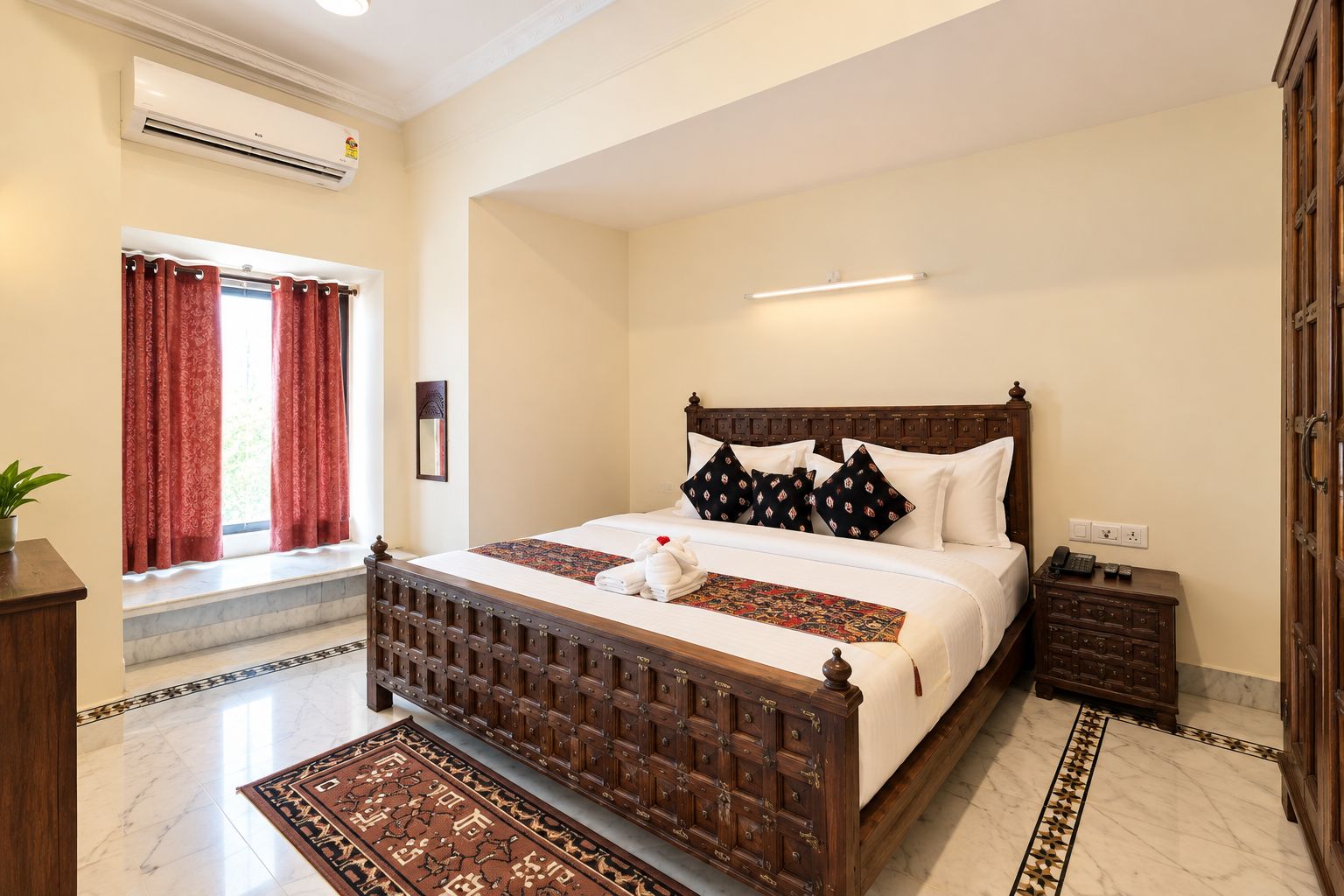 A hotel room with a large bed, a wooden headboard, a bedside table, a nightstand, a lamp, a window with curtains, and a wall-mounted air conditioning unit.