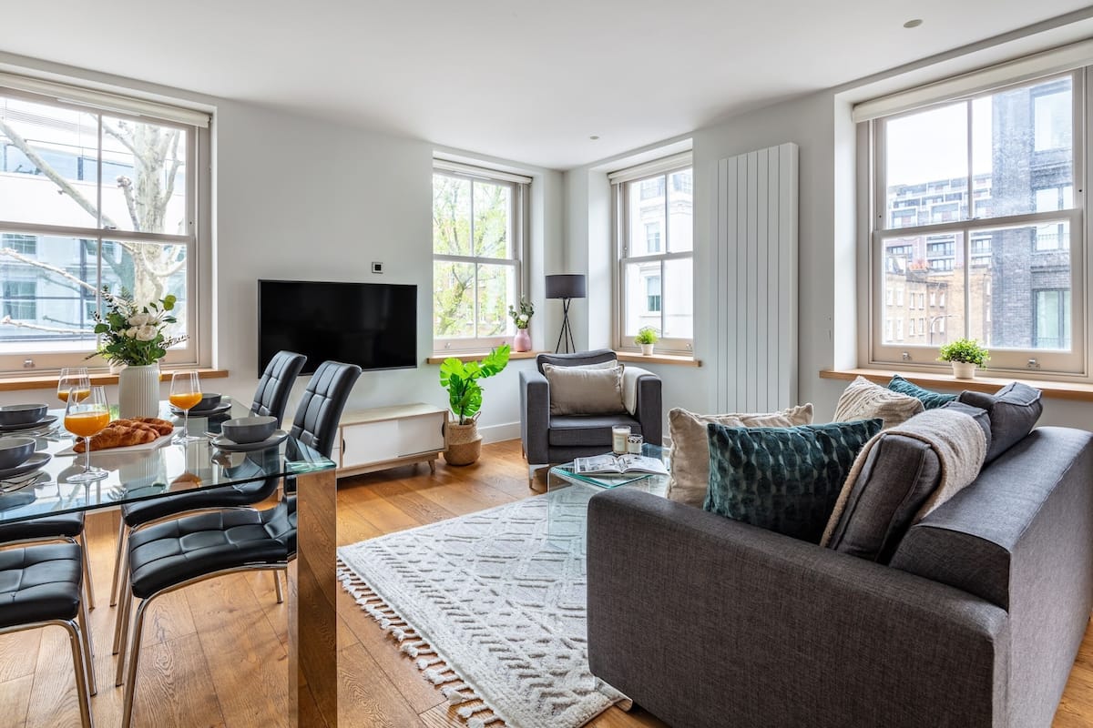 Una sala de estar moderna con una mesa de comedor de vidrio, sillas de cuero negro, un sofá gris y ventanas amplias que ofrecen vistas de la ciudad.