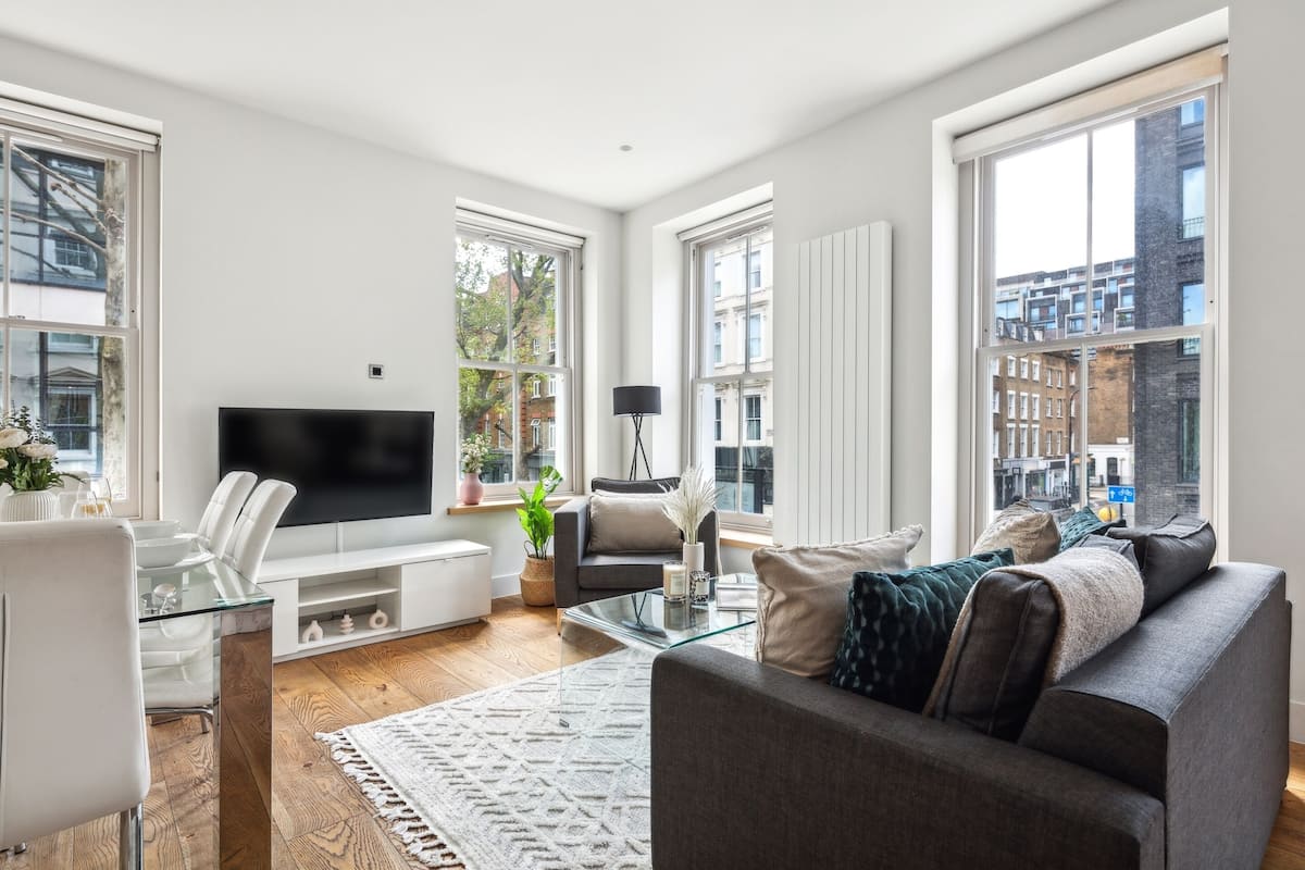 Una sala de estar moderna con un televisor de pantalla plana grande, una mesa de centro de vidrio, un sofá gris y una mesa de comedor blanca con sillas.