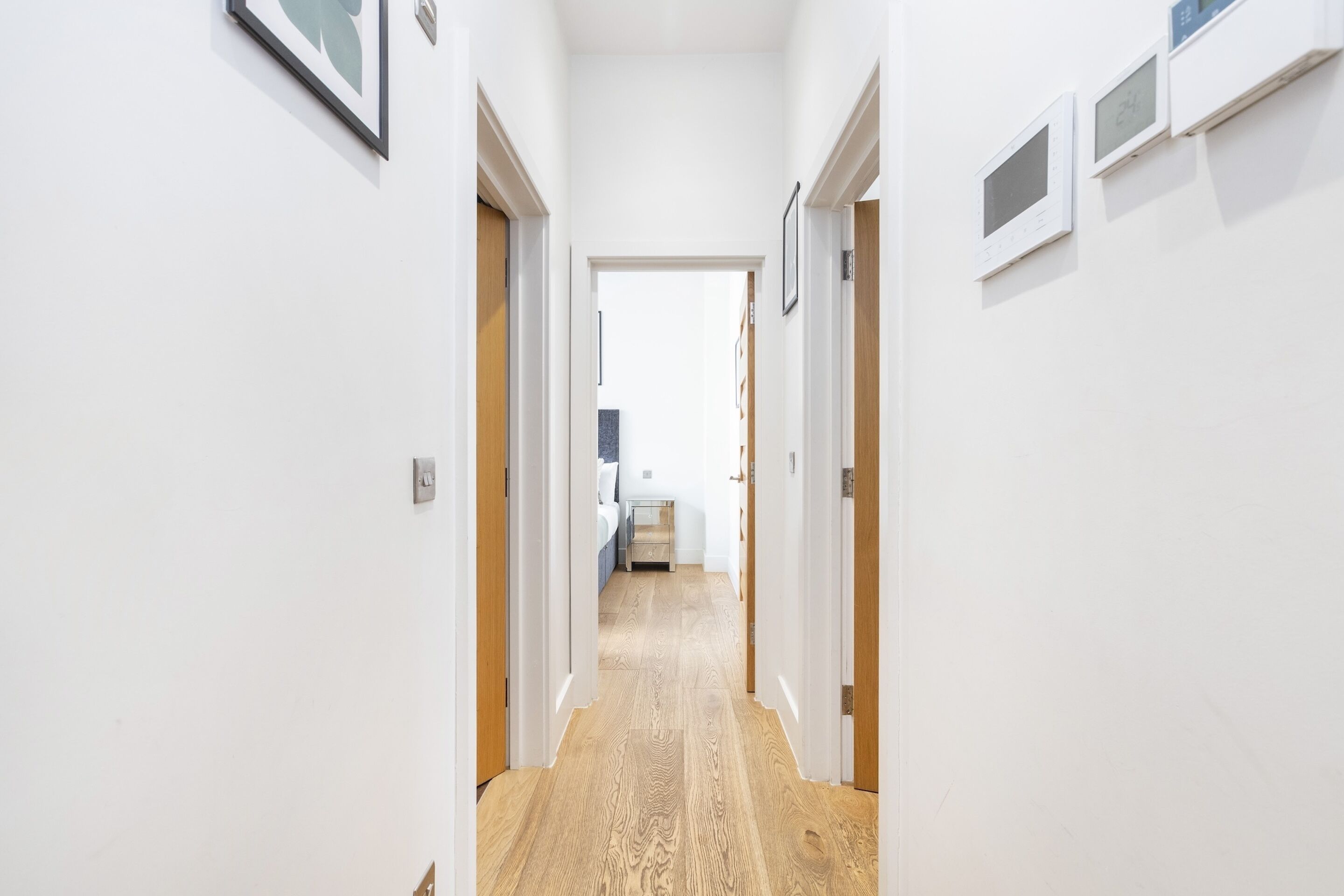 Un pasillo estrecho con suelo de madera y paredes blancas. Hay cuadros en las paredes y una puerta al final que da a un dormitorio.