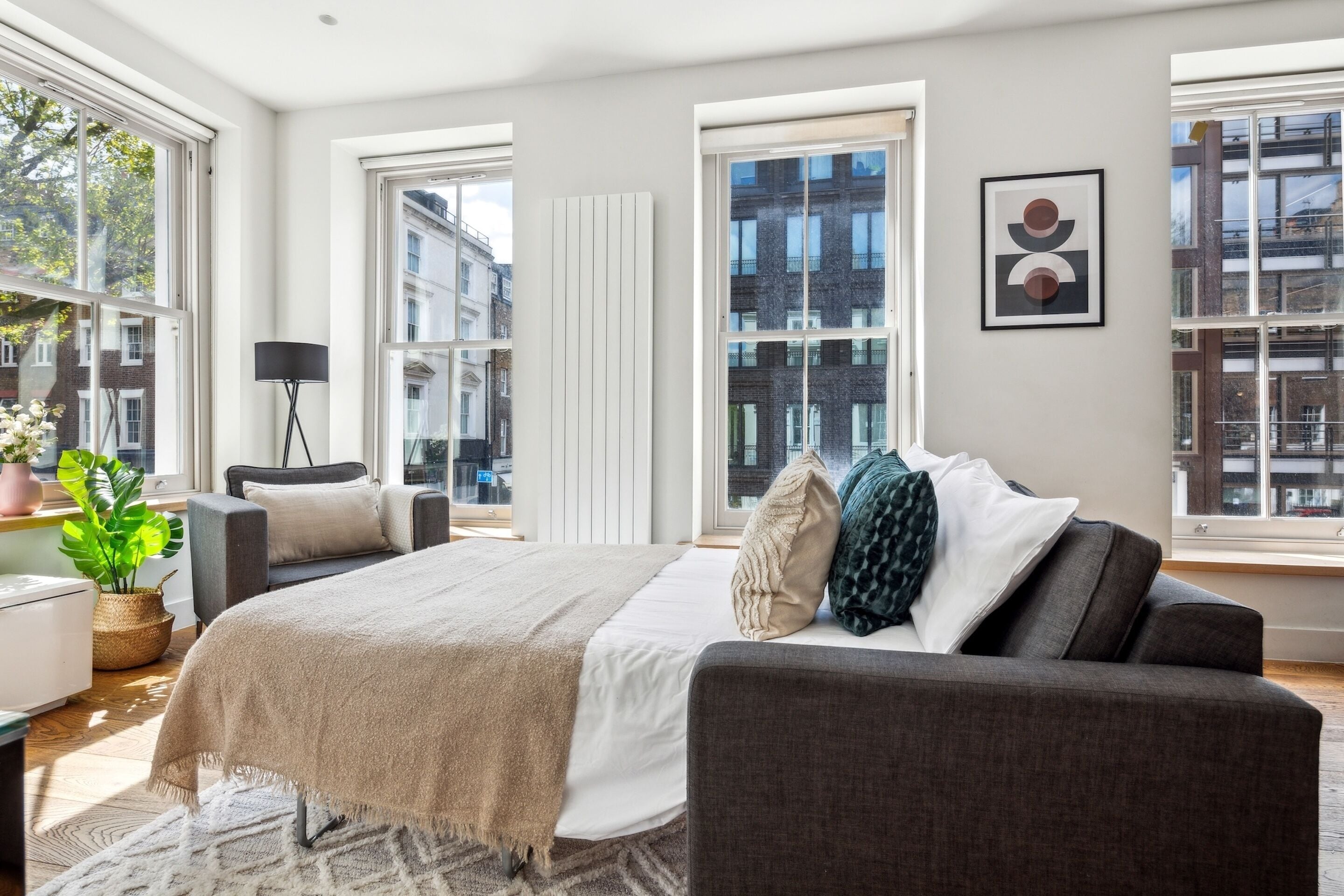 Ein modernes Schlafzimmer mit einem großen Bett, einem Nachttisch, einer Lampe und Blick auf die Stadt durch große Fenster.
