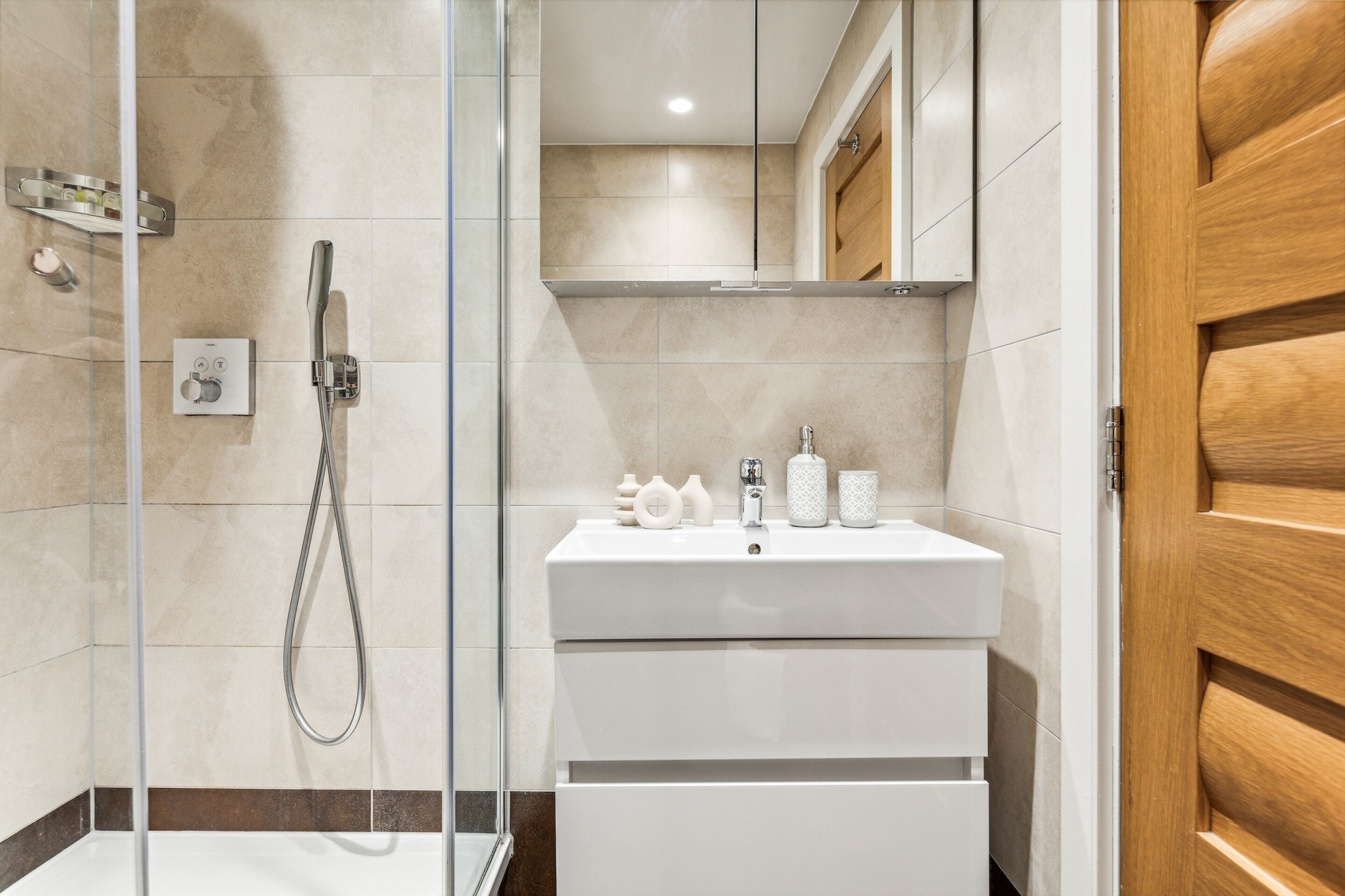A modern bathroom with a glass-enclosed shower, a white sink with a mirror above, and a wooden door.