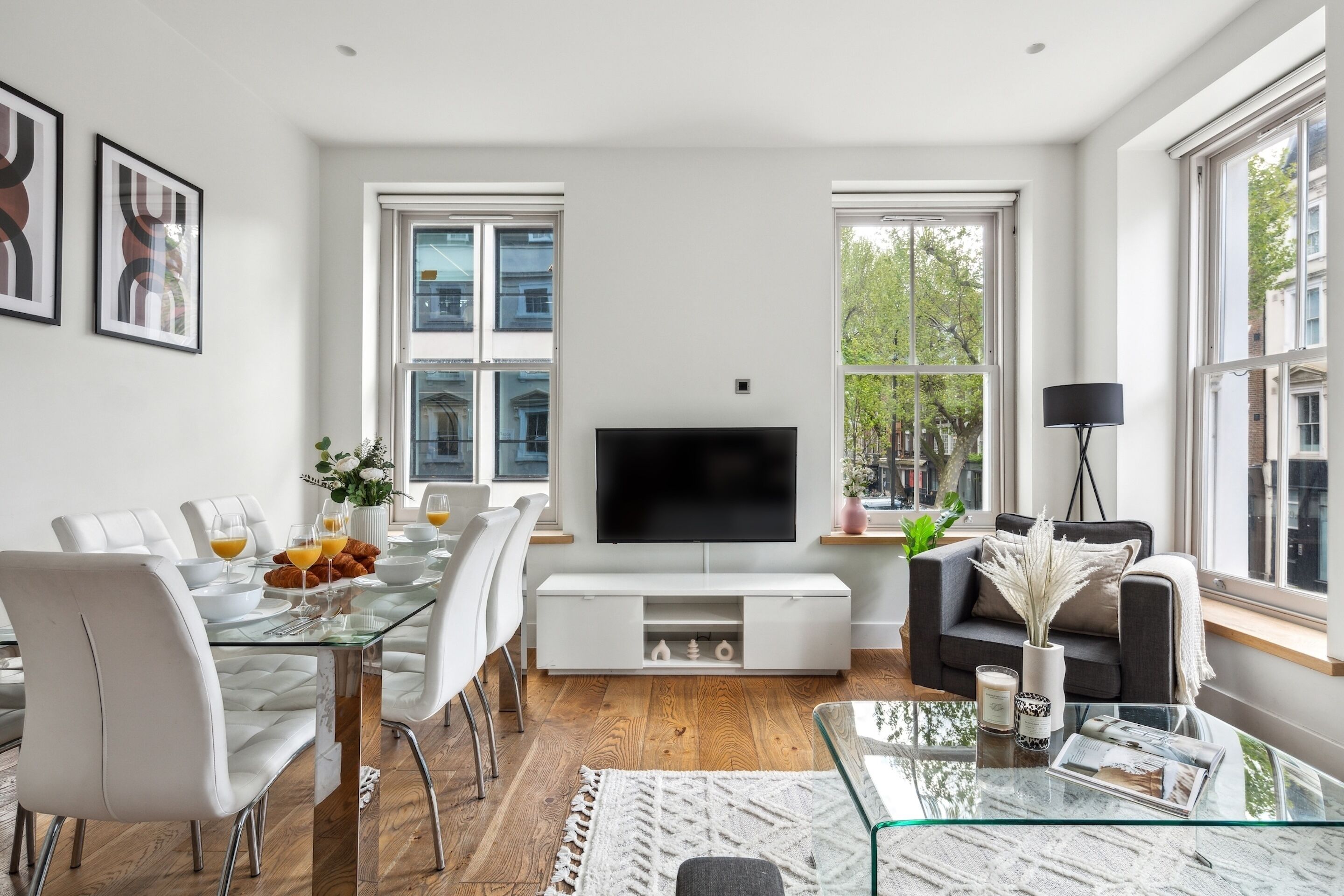 A modern living room with a glass dining table, white chairs, a flat-screen TV, a sofa, and large windows.
