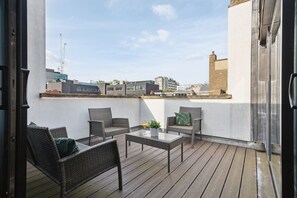 Un balcon doté de meubles en osier et d’une table en verre, offrant une vue sur le paysage urbain avec ses bâtiments et sa grue.