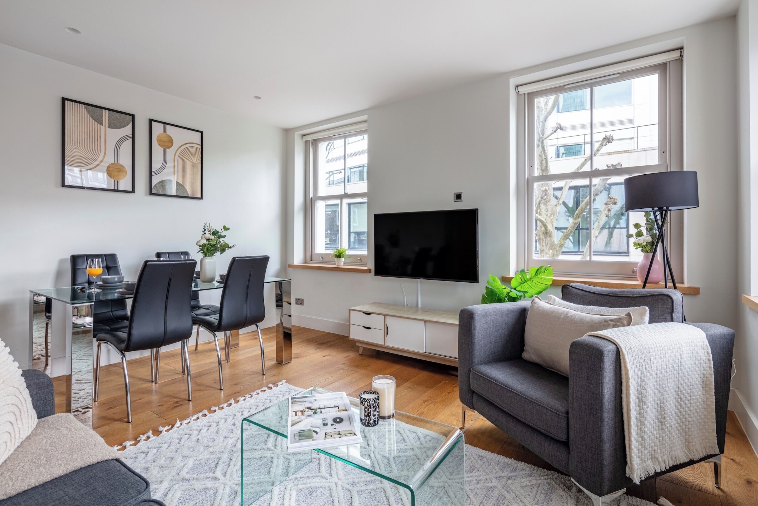 En moderne stue med spiseplads, store vinduer, et fladskærms-tv og en grå sofa.
