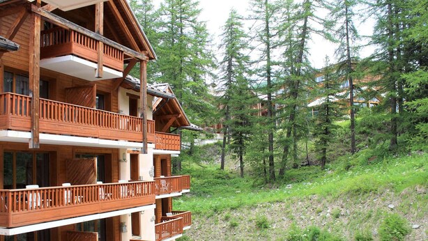 Un immeuble en bois à plusieurs étages, doté de balcons, entouré d’arbres et situé au pied d’une colline.