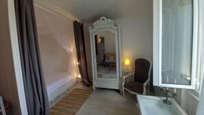 Une chambre d’hôtel avec un lit, une chaise, une petite table, un miroir et une fenêtre avec des rideaux.