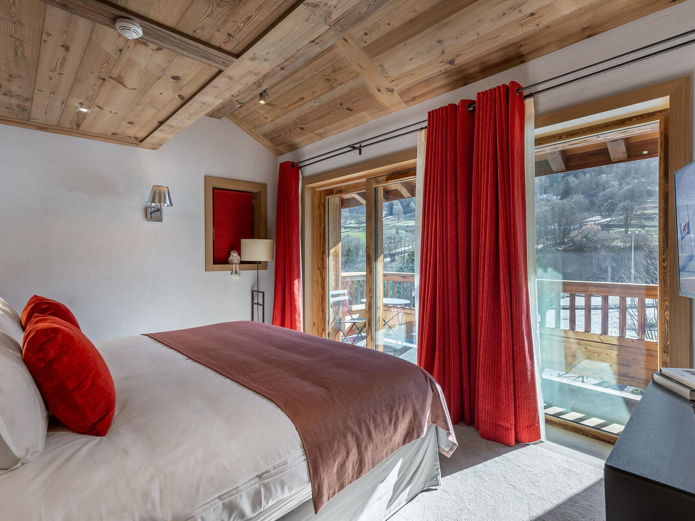 A bedroom with a wooden ceiling, a bed with white and red bedding, a nightstand, a lamp, and a view of a balcony with red curtains.