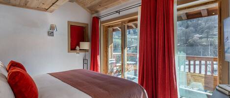 A bedroom with a wooden ceiling, a bed with white and red bedding, a nightstand, a lamp, and a view of a balcony with red curtains.