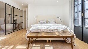 Un dormitorio con una cama grande, un banco de madera, una bañera y vistas de la ciudad a través de una ventana.
