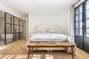 Un dormitorio con una cama grande, un banco de madera, una bañera y vistas de la ciudad a través de una ventana.