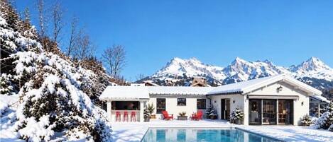 Paisaje nevado con piscina, una casa con patio y montañas al fondo.