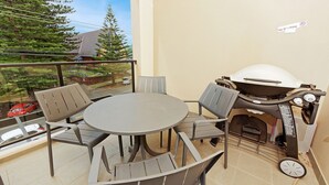 Ein Balkon mit rundem Tisch und vier Stühlen, einem Grill und Blick auf eine Wohngegend mit Bäumen und einem Auto.