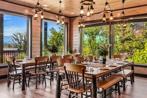 A dining area with wooden tables and chairs, large windows with a view of greenery, and hanging lantern-style lights.