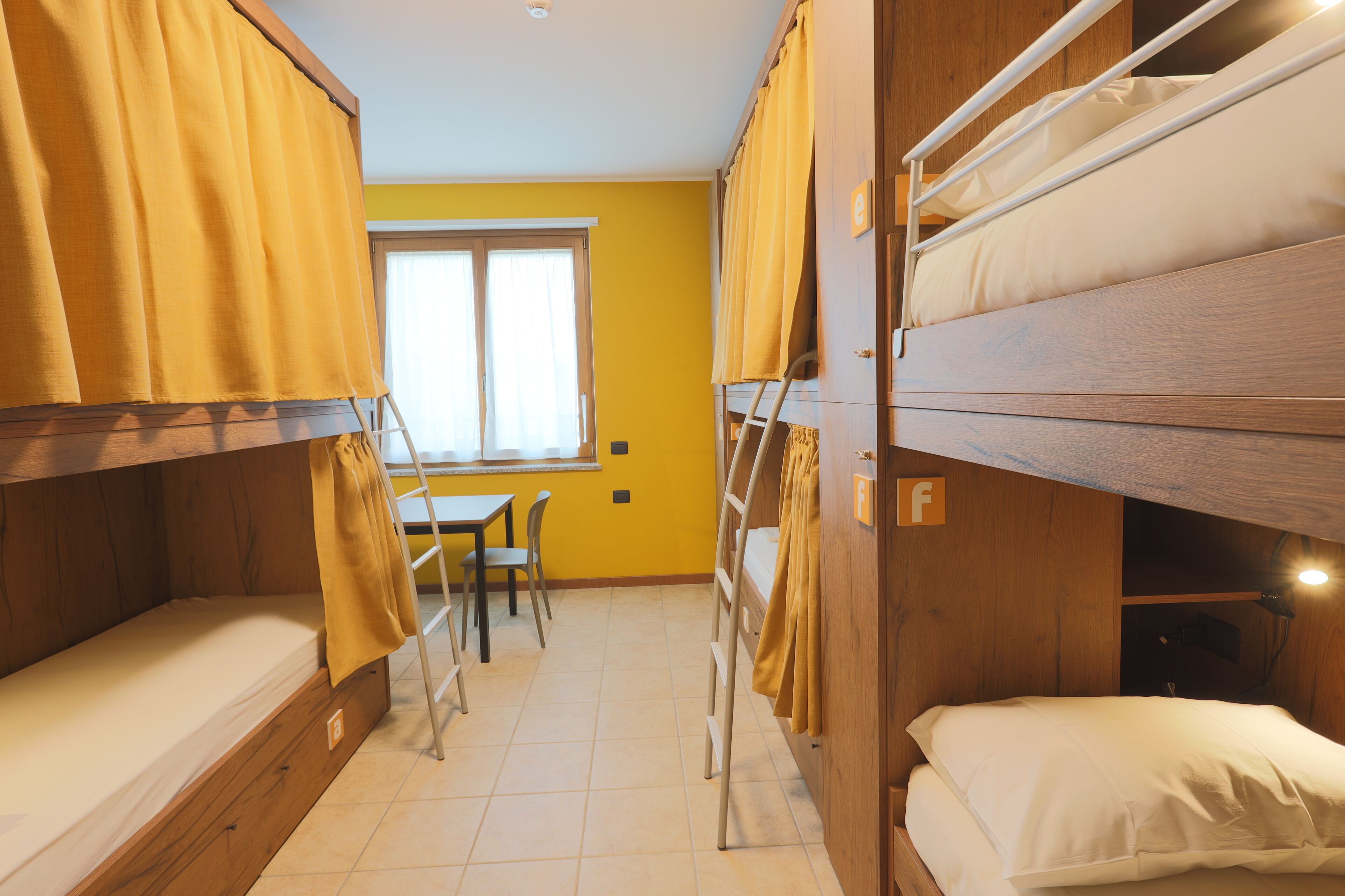 Um quarto de dormitório com beliches, uma mesa, cadeiras e uma janela.
