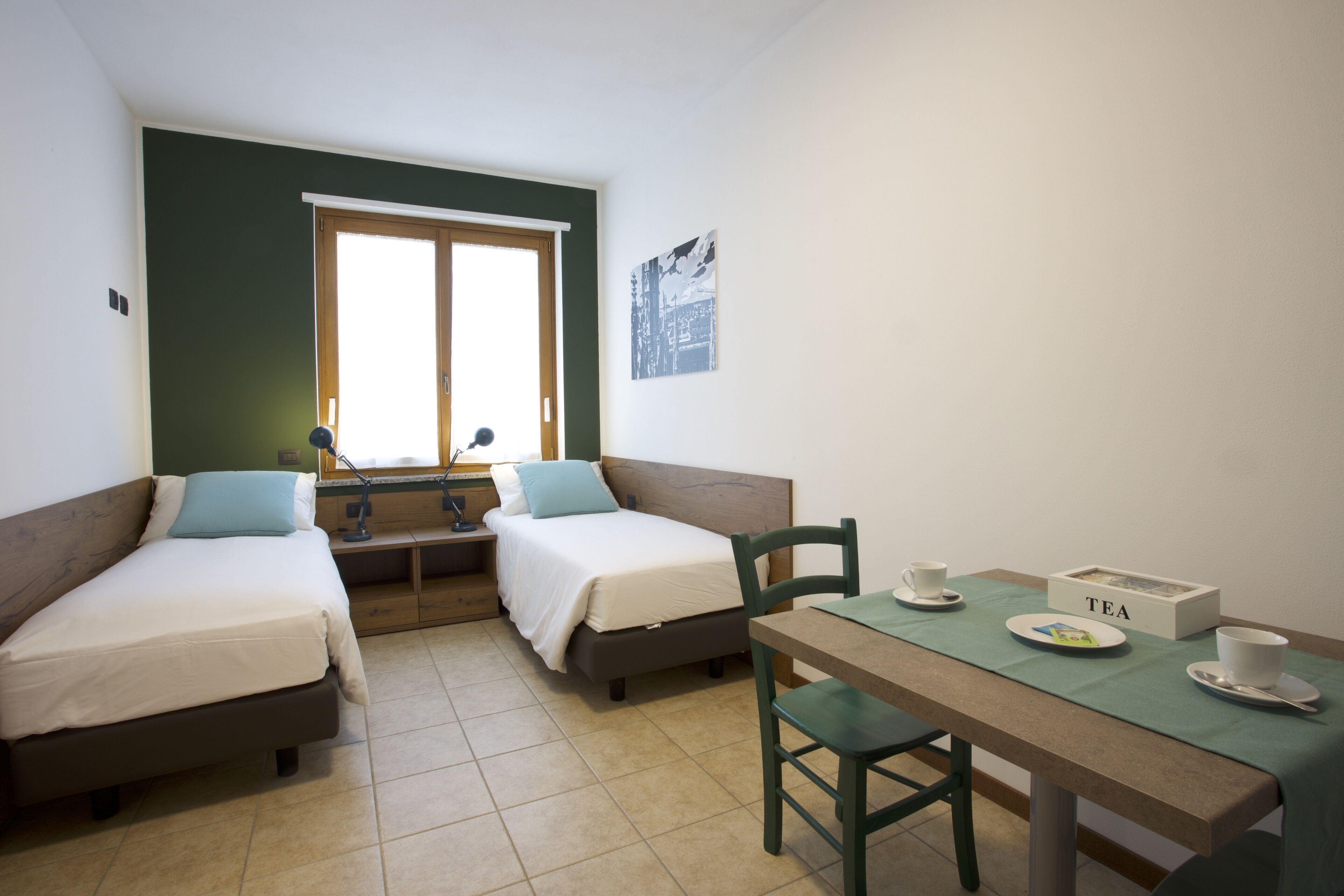 A small, well-lit room with two single beds, a table with a tea set, and a window with a view.