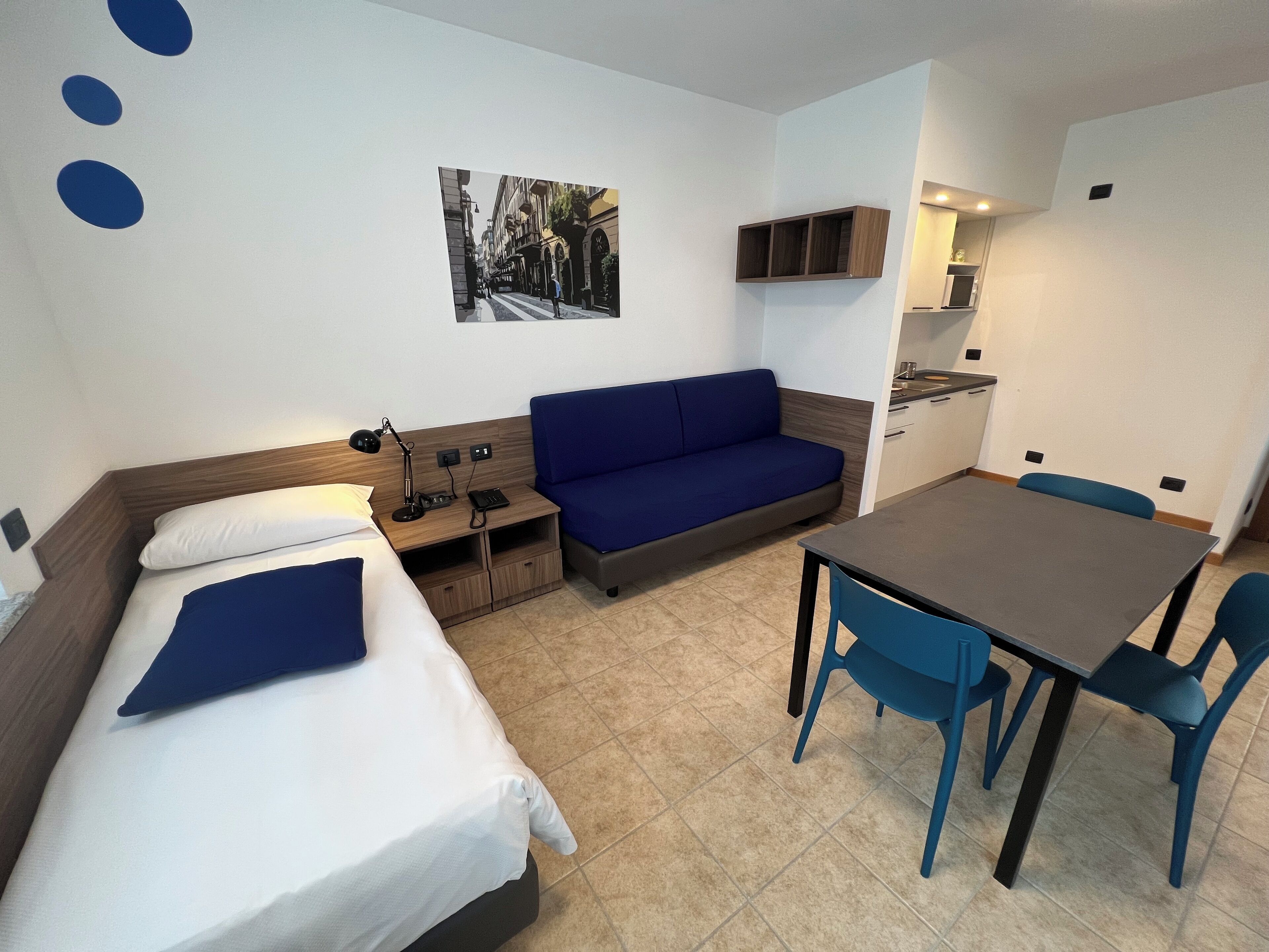 A compact hotel room with a bed, a sofa, a small table, and a kitchenette.
