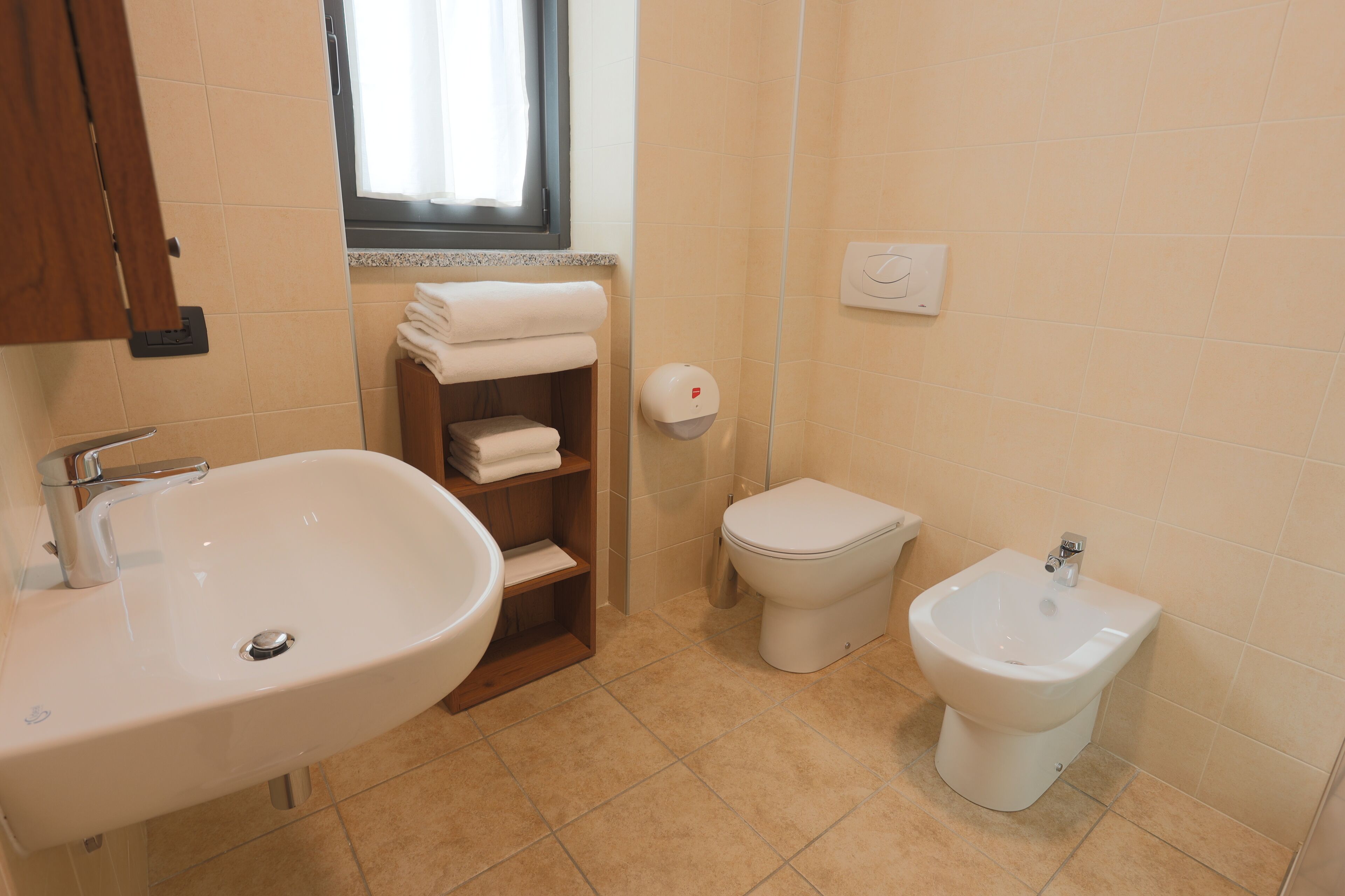 A bathroom with a sink, toilet, bidet, and a shelf with towels.