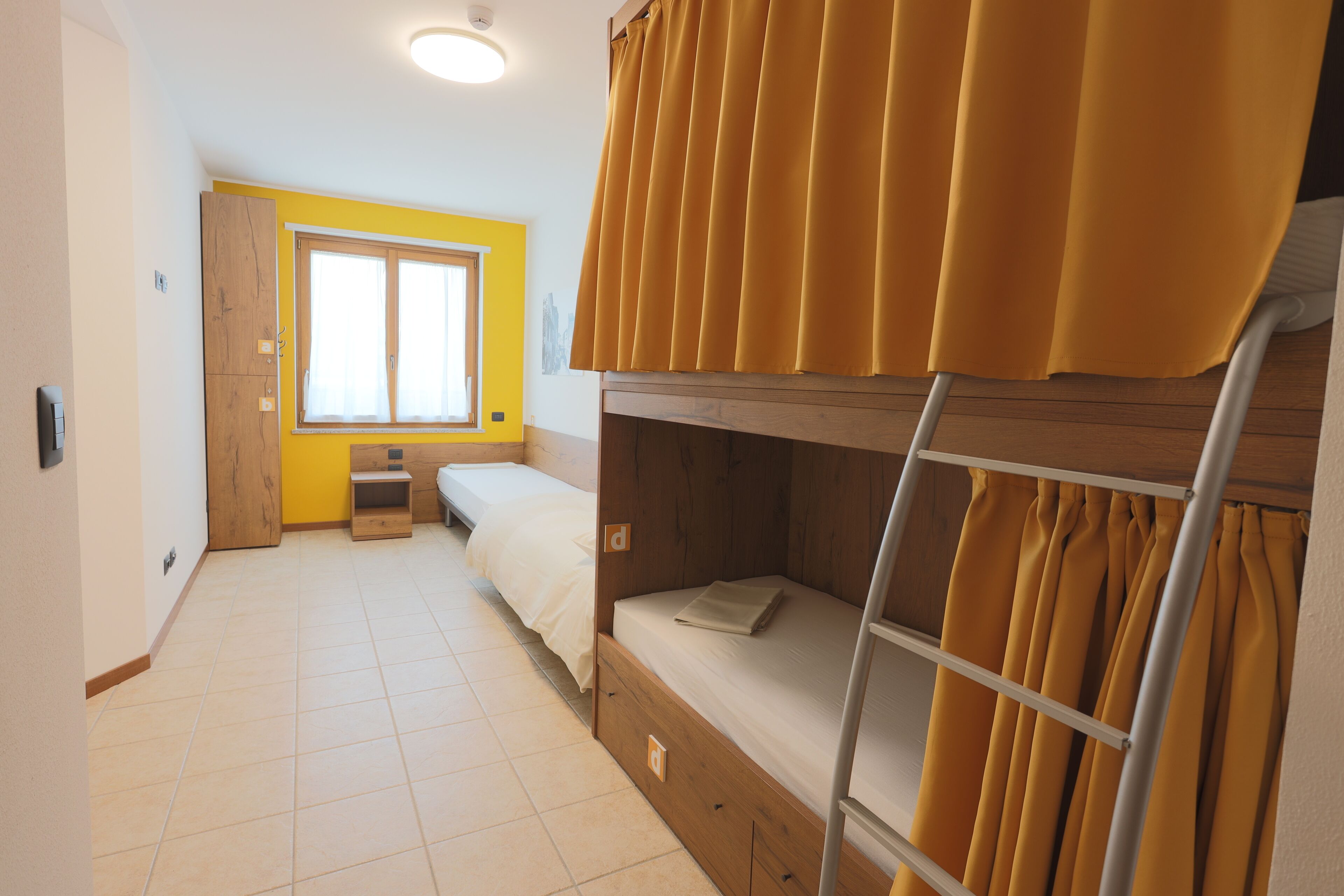 A dormitory room with a bunk bed, a desk, a chair, a window with curtains, and a door.