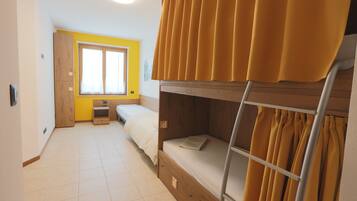 A dormitory room with a bunk bed, a desk, a chair, a window with curtains, and a door.