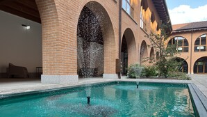 A swimming pool with water jets, surrounded by brick arches and a building with large windows. - Corte Milano - Hostel (Rozzano)