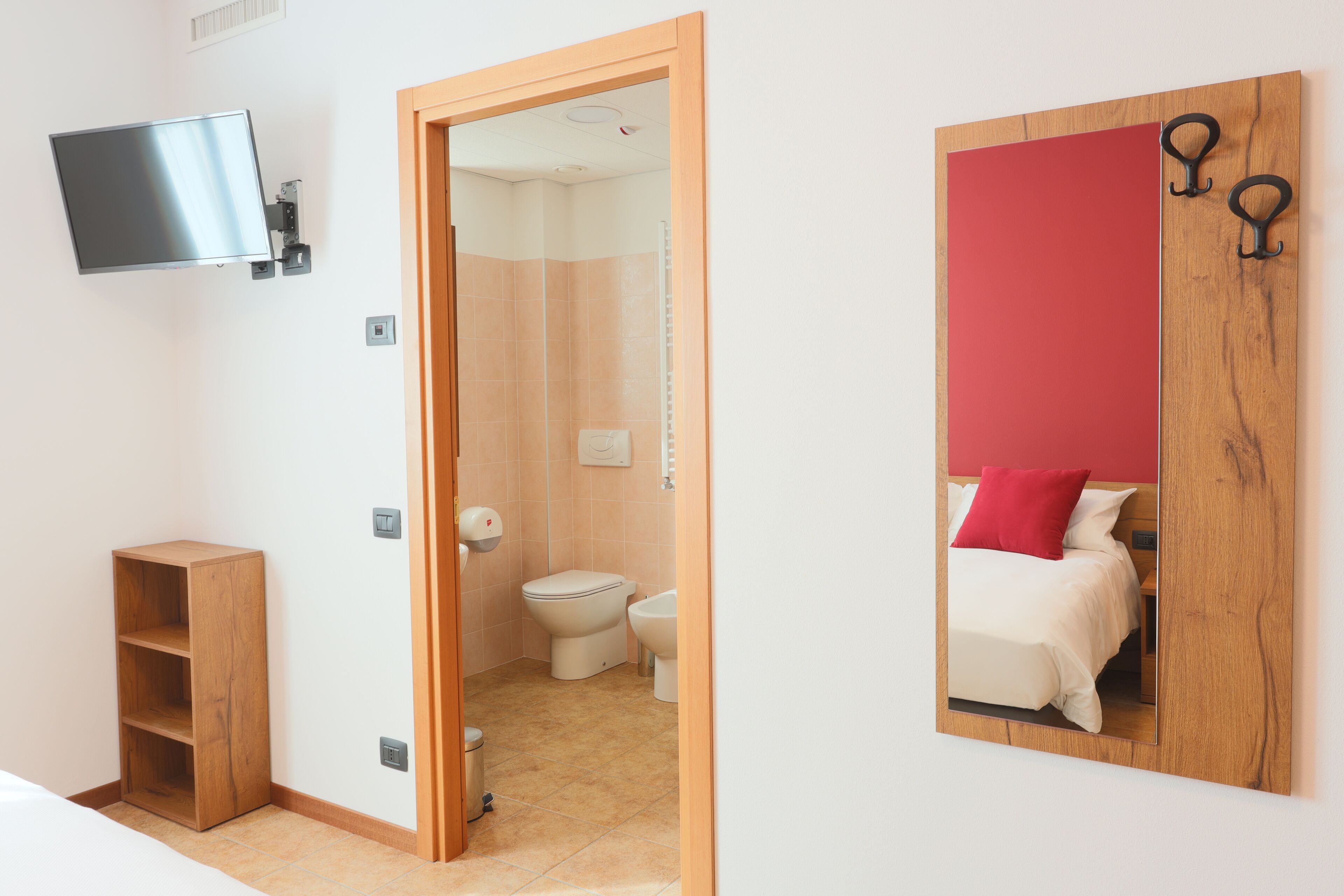 Habitación de hotel con televisor montado, estante de madera, armario de madera y espejo con panel rojo.