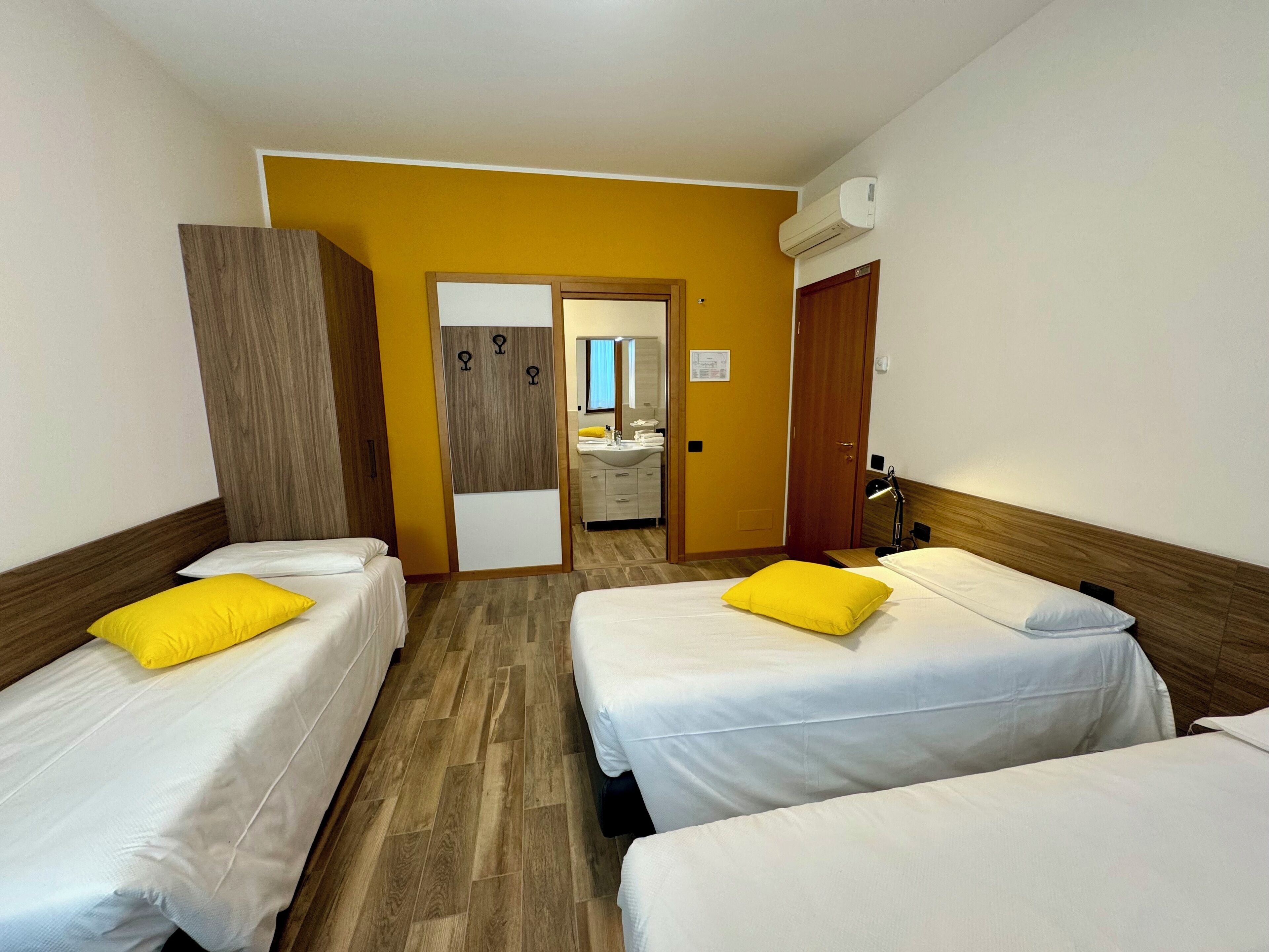 A hotel room with two single beds, a wooden wardrobe, a mirror, and a bathroom visible through an open door.