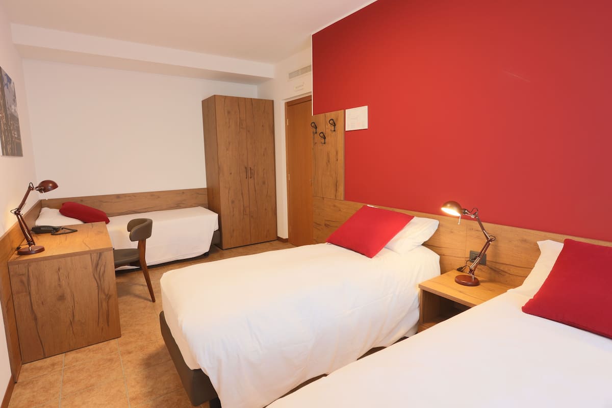 A hotel room with two beds, a wooden wardrobe, a desk with a lamp, and a red accent wall.