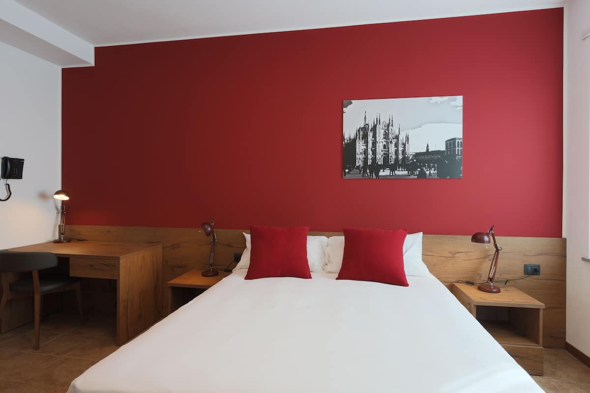 A hotel room with a red accent wall, a bed with white bedding, two red pillows, a wooden desk, a chair, a nightstand with a lamp, and a framed picture on the wall.