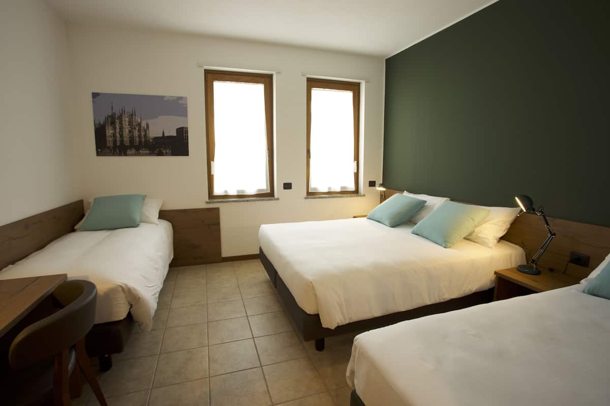 A room with two single beds, a desk, and a chair. There are two windows providing natural light.