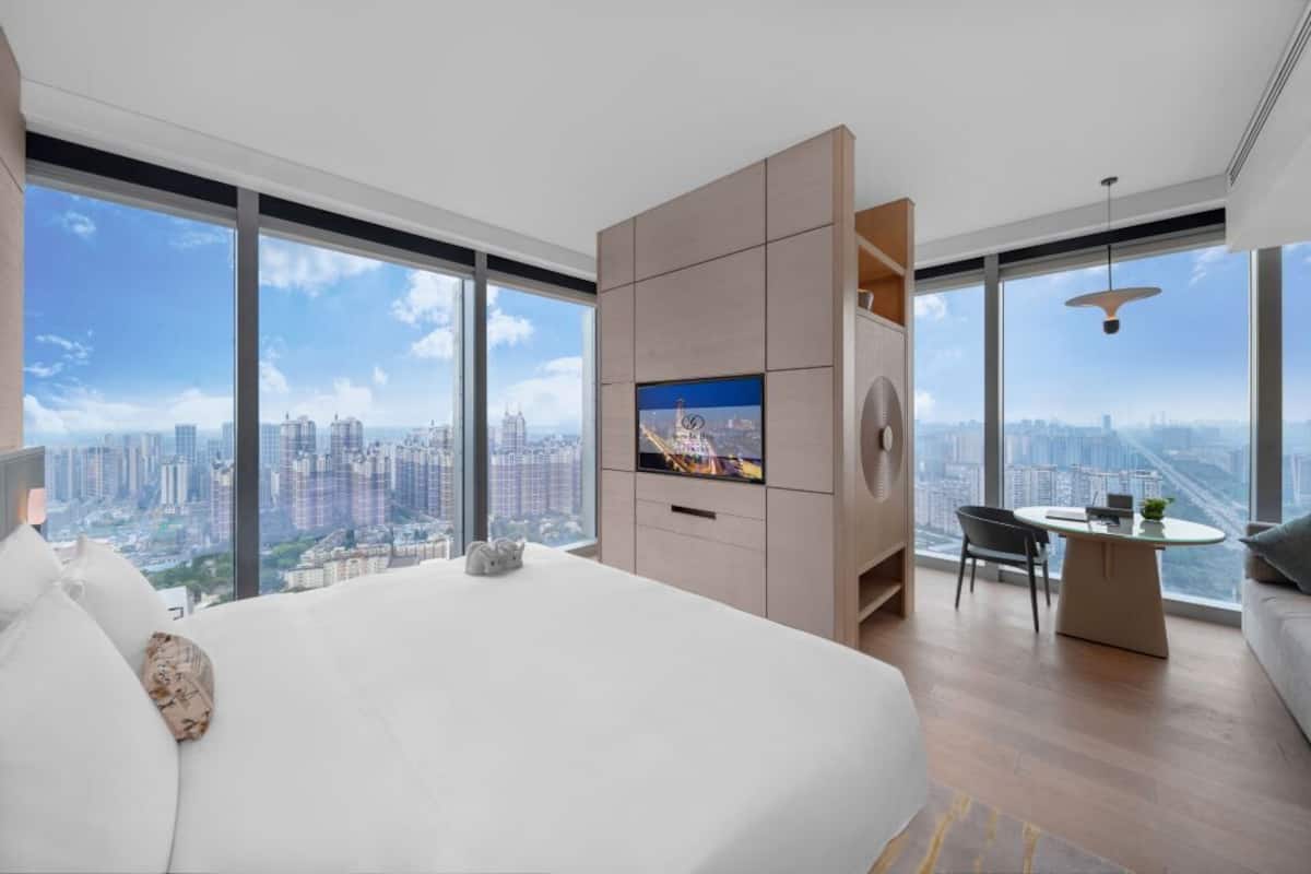A modern hotel room with a large bed, a TV, a desk, and a view of a cityscape.