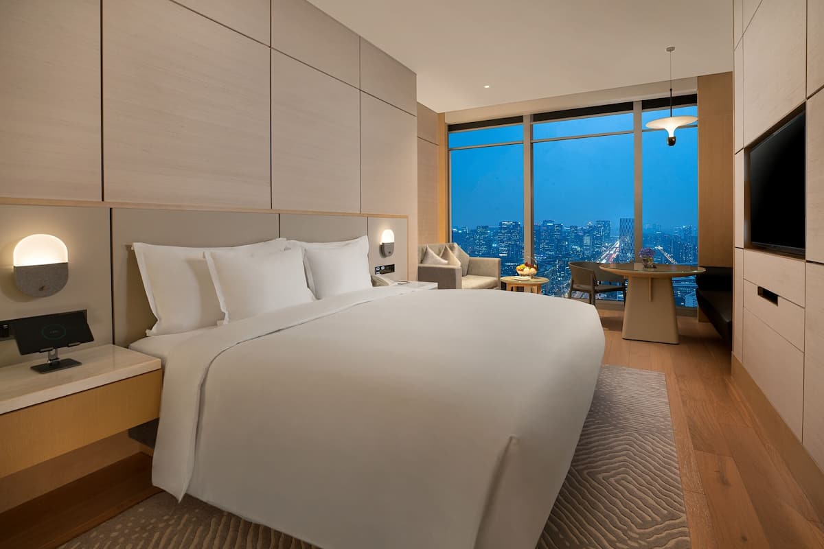 A modern hotel room with a large bed, a desk, a chair, a TV, and a view of the cityscape.