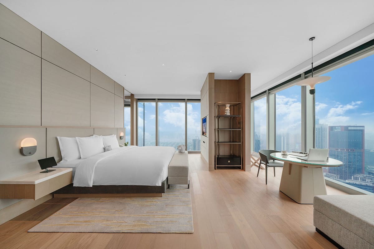 A modern bedroom with a large bed, bedside tables, a desk, and a view of the city.