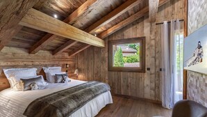 Ein Schlafzimmer einer Holzhütte mit einem Bett, Kissen, einem Sessel und einem Fenster mit Blick auf Grünflächen.