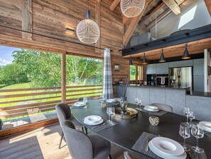 Un comedor moderno con una gran mesa dispuesta para seis personas, rodeado de vigas de madera y amplias ventanas que dan a un jardín exuberante.