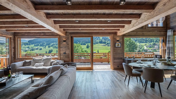 Ein geräumiger Raum mit Holzbalkendecke, großen Fenstern mit Blick auf eine grüne Landschaft, einem Sofa und einem Essbereich mit Stühlen.