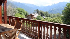 Una terraza de madera con una mesa y sillas, con vista a un paisaje montañoso donde se ve una cabaña a lo lejos.