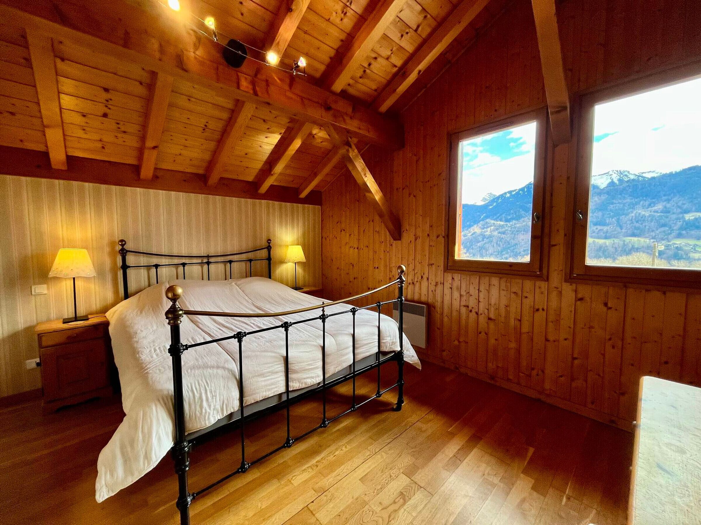 A wooden cabin bedroom with a metal bed frame, two bedside lamps, and a window with a mountain view.