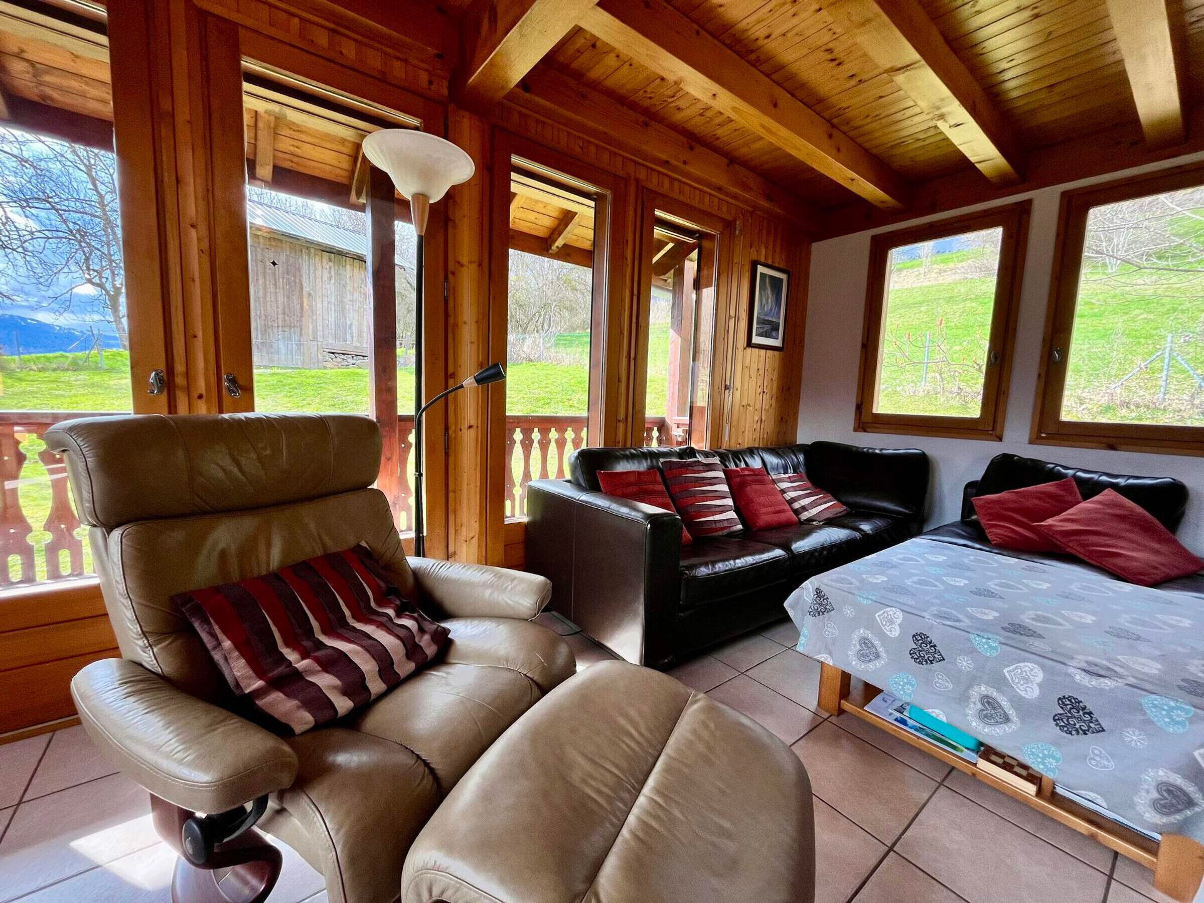 A cozy room with a leather chair, a black sofa, and a bed with patterned bedding. Large windows offer a view of a green landscape.