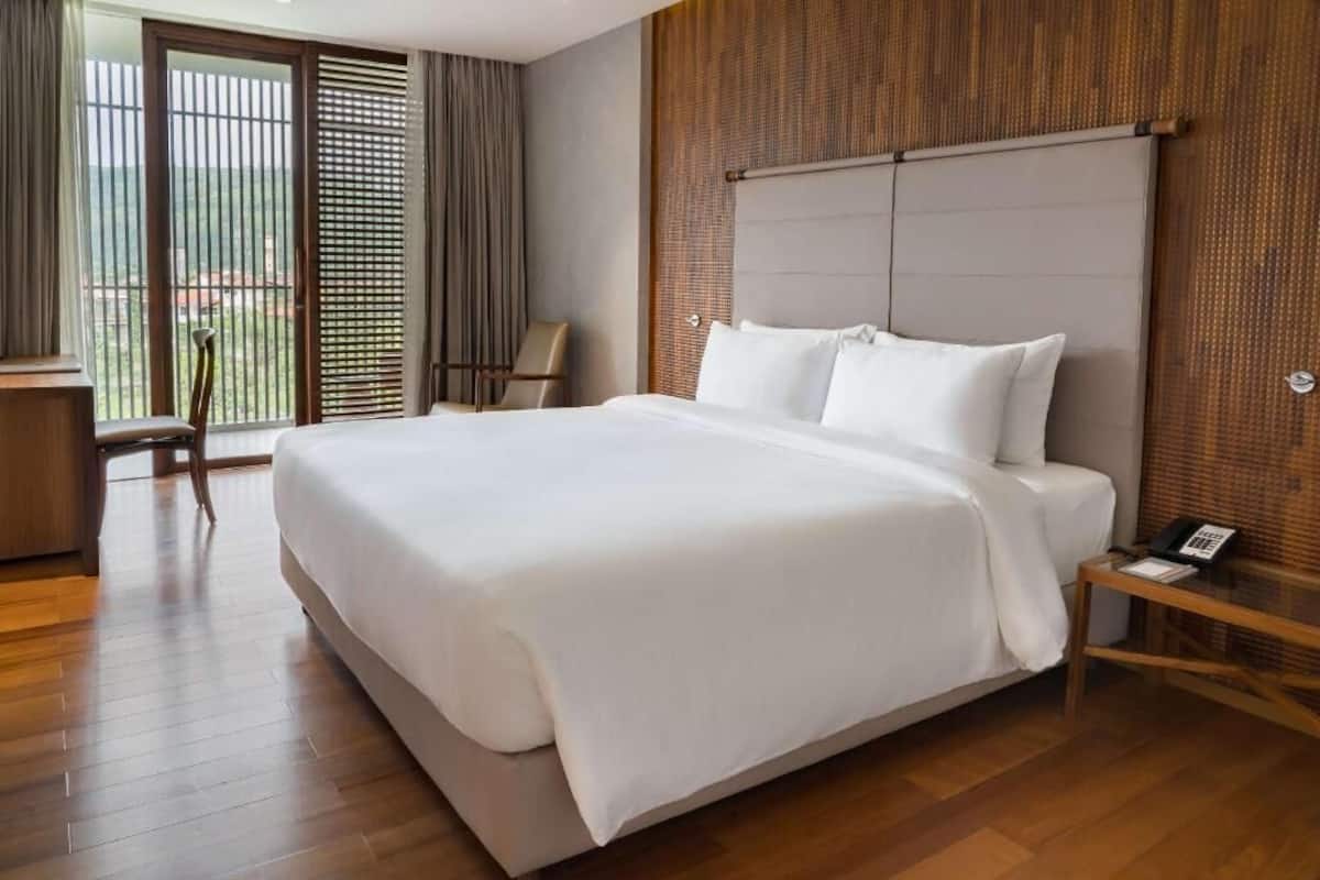 A hotel room with a large bed, a desk with a chair, a small table with a phone, and a view through a sliding door.