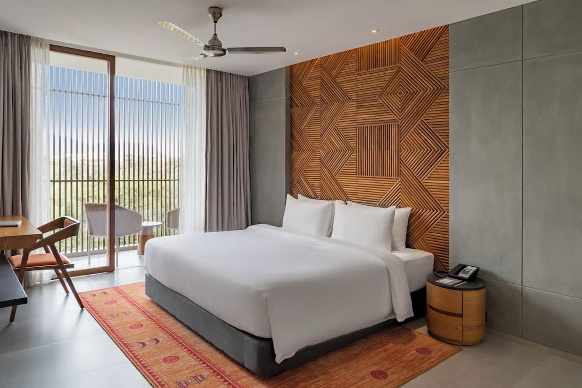 A modern hotel room with a large bed, a desk, and a chair, a balcony with curtains, and a patterned wall.