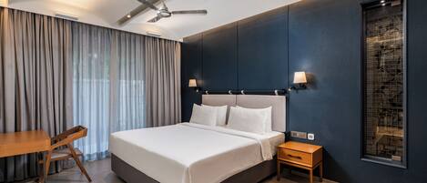 Una habitación de hotel moderna con una cama grande, un escritorio, una silla, una mesita de noche, un ventilador de techo y una ventana con vistas.