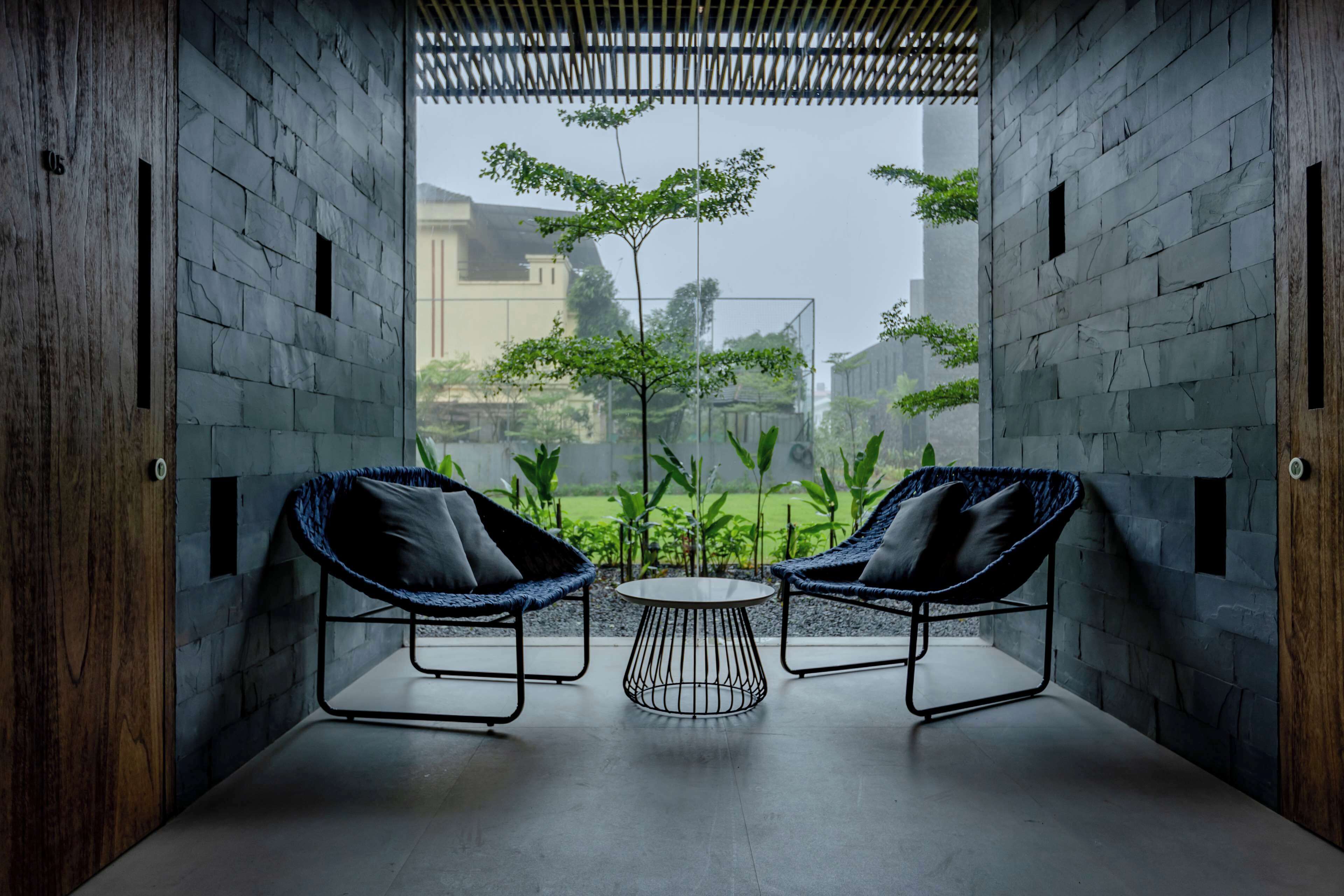 A modern interior with two wicker chairs and a small round table, set against a backdrop of a brick wall and a view of greenery outside.