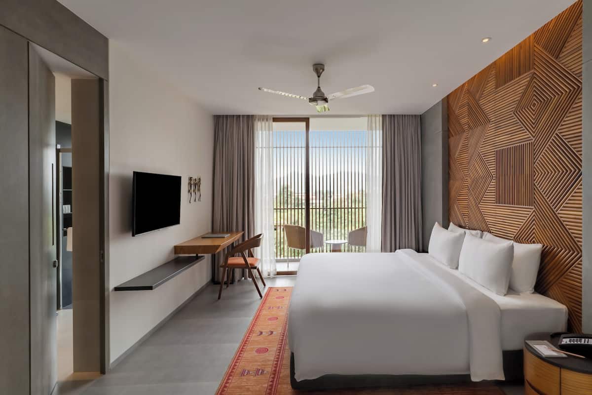 A modern hotel room with a large bed, a desk, a chair, a TV, and a balcony with curtains.