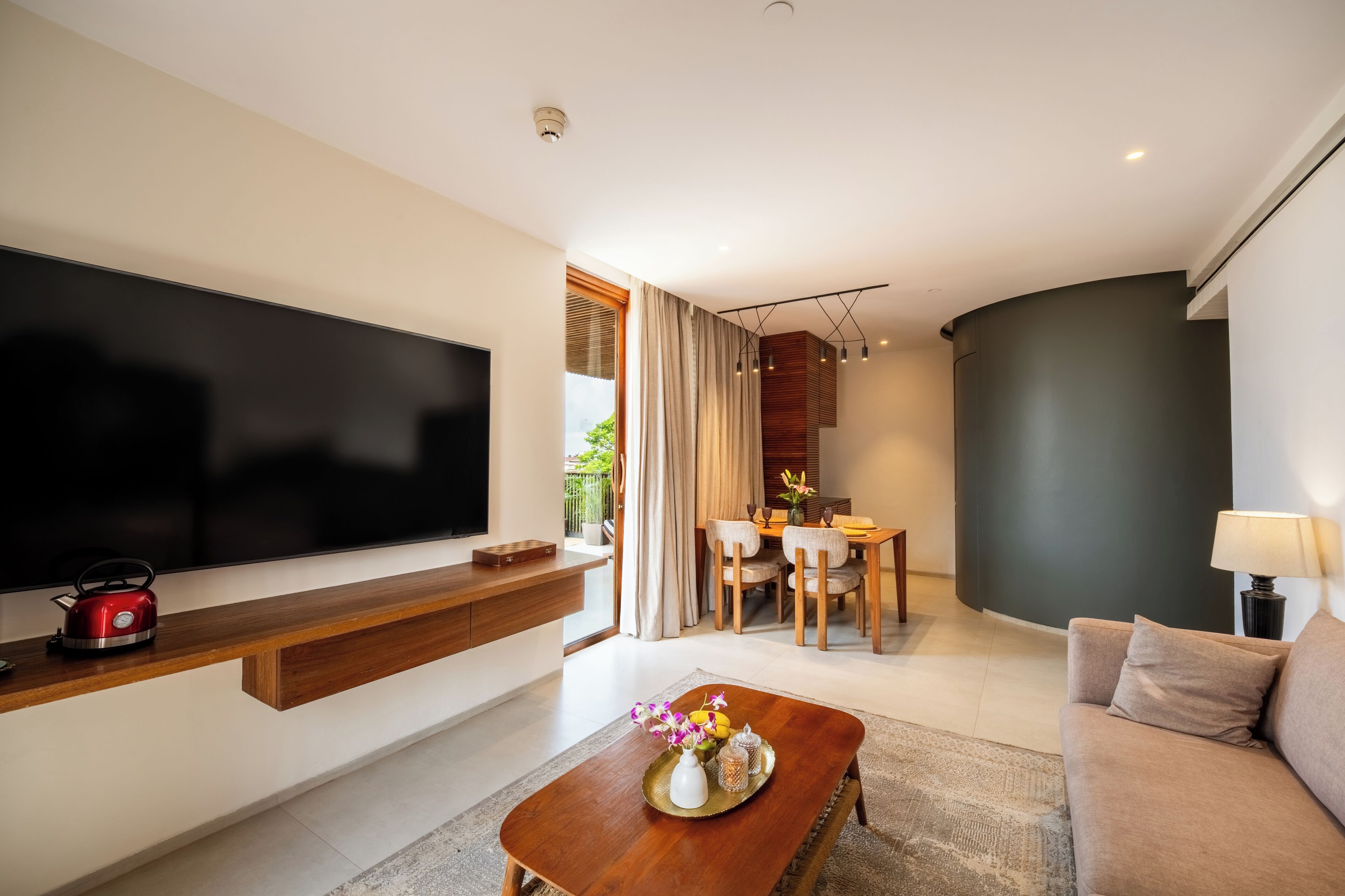 A modern living room with a flat-screen TV, a wooden coffee table, a sofa, a dining area with a table and chairs, and a large window with curtains.