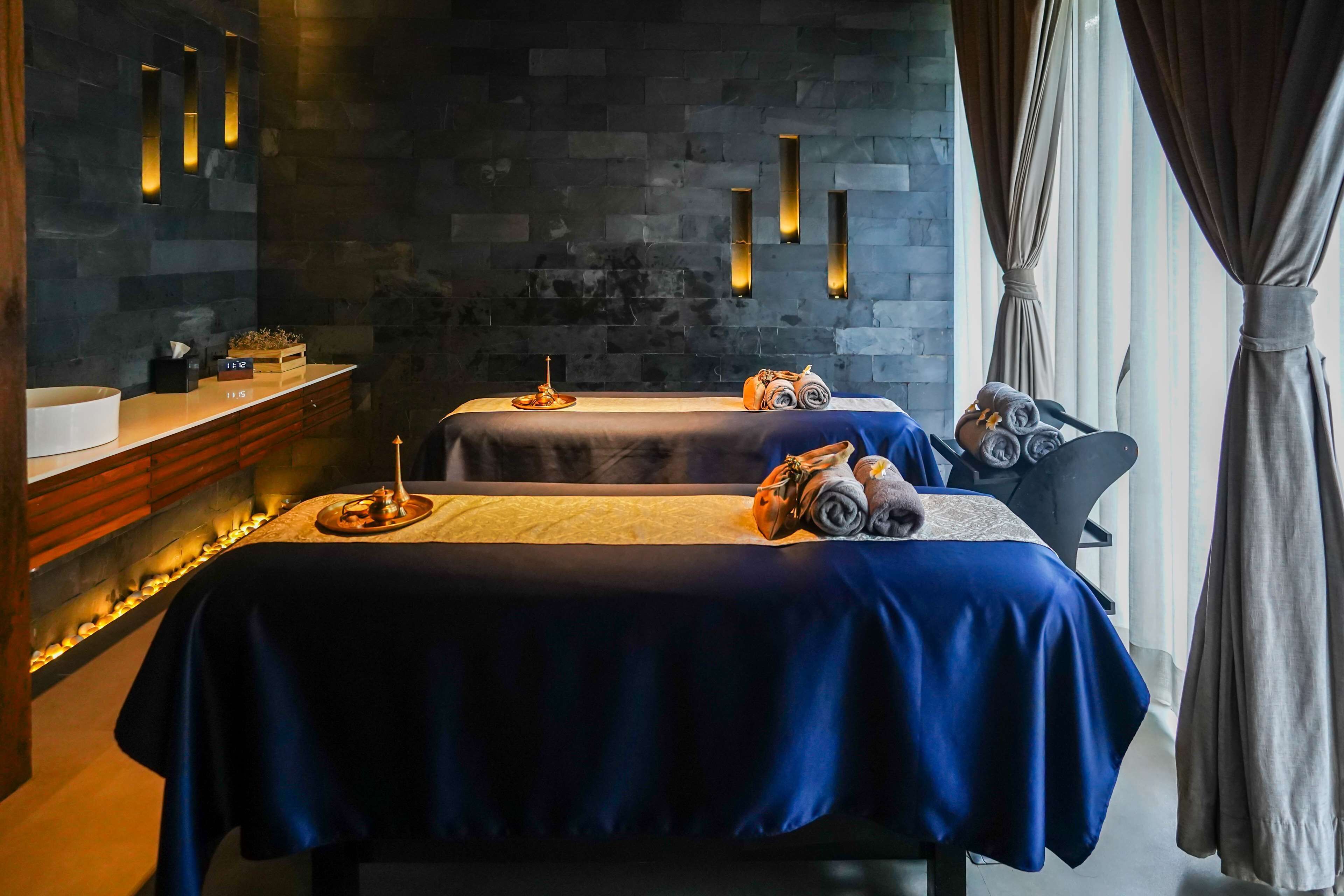 Un cuarto de spa con dos camillas de masaje, un banco de madera, un pequeño altar y una pared de piedra con iluminación empotrada.