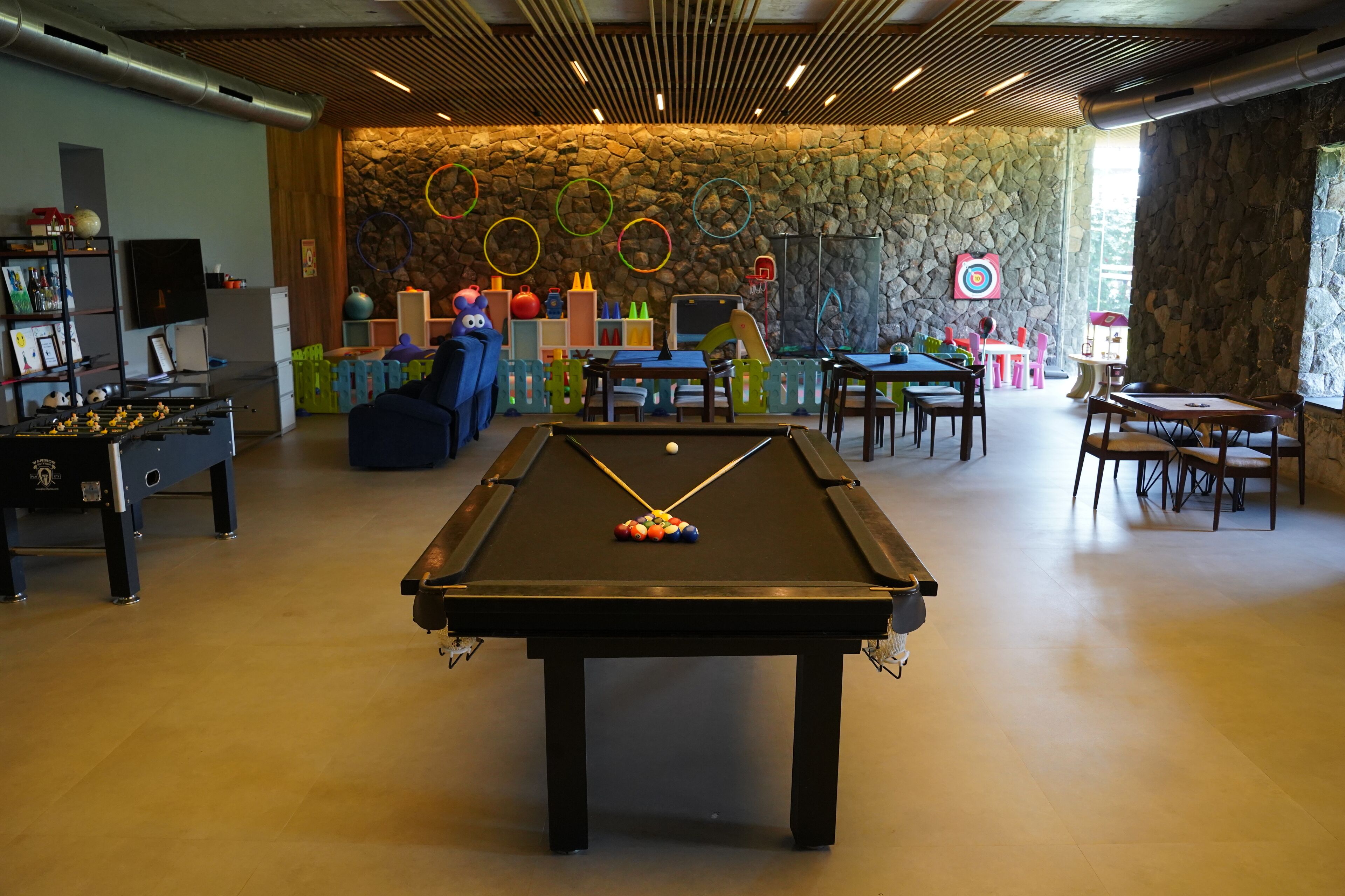 A pool table with colorful balls, a foosball table, and a dartboard in a room with stone walls and a wooden ceiling.