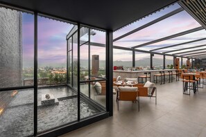 Un bar moderno en la azotea con paredes de vidrio, asientos al aire libre y vistas de la ciudad.