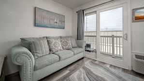 A living room with a light green sofa, patterned cushions, a painting of sailboats, a large window with curtains, and a view of the sea. - New Listing!  Modern studio ocean block with a pool (Ocean City)